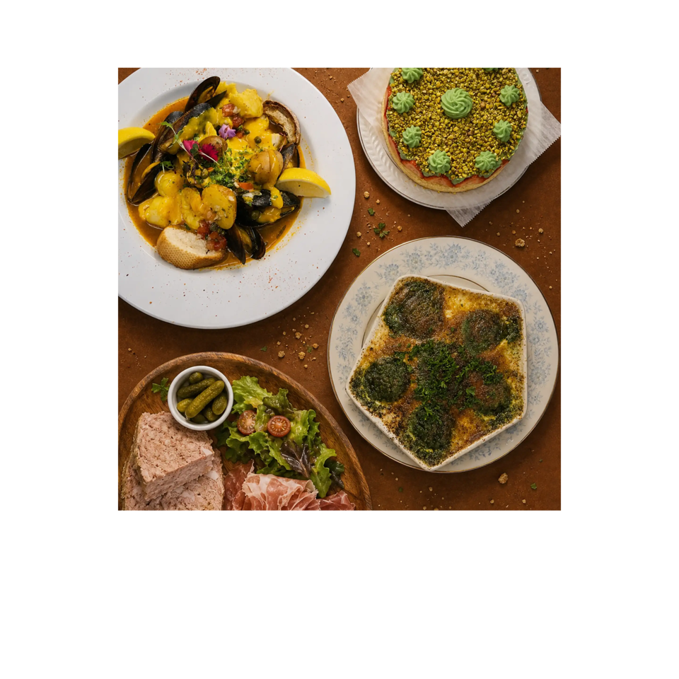 Overhead view of a restaurant spread featuring seafood stew, pistachio cake, herb-topped baked dish, charcuterie, salad, and pickles.
