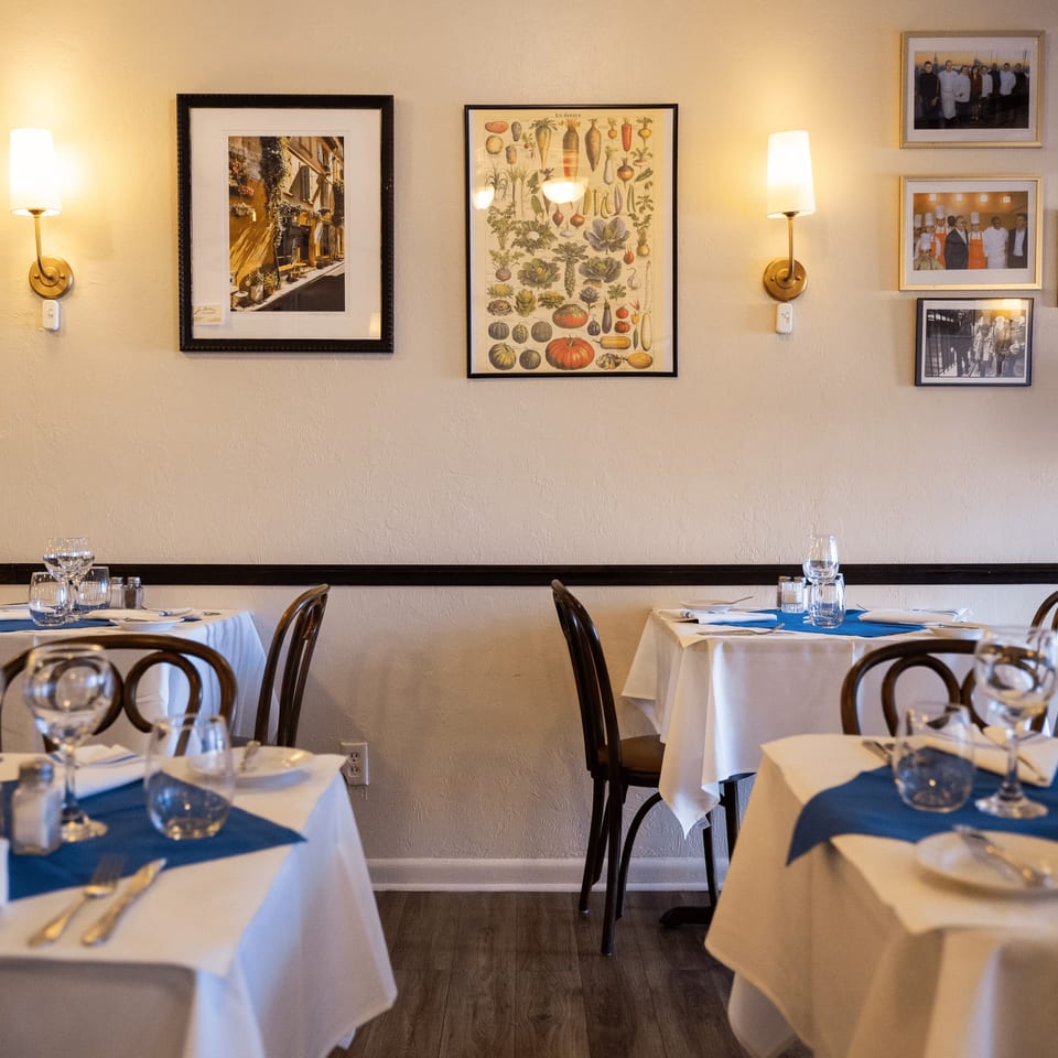 Warmly lit restaurant dining room with white tablecloths, blue napkins, bentwood chairs, framed artwork, and wall sconces.